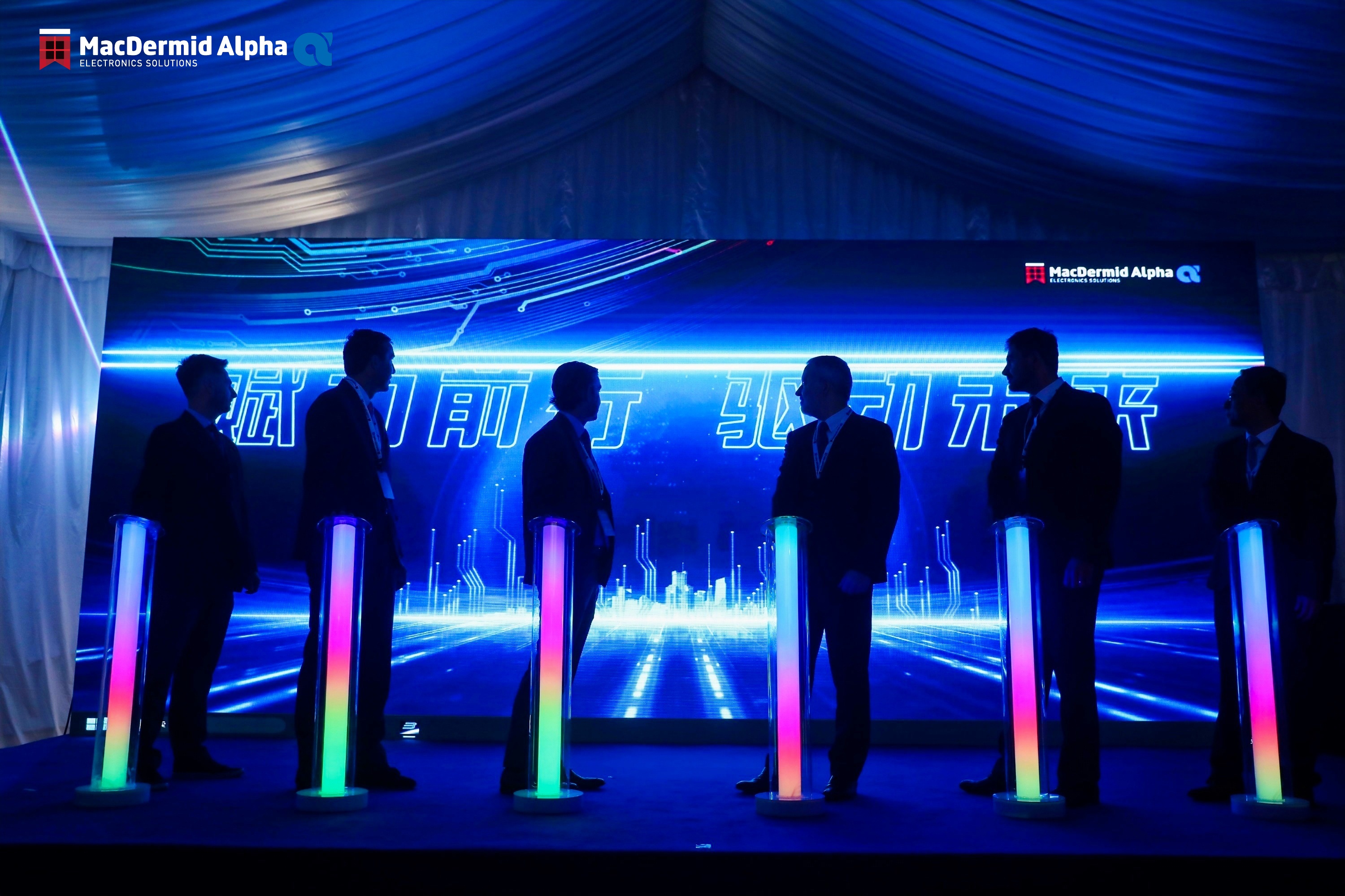 macdermid alpha business men are darkened silhouettes standing at rainbow-LED podiums in a dark room with neon blue backdrop on stage